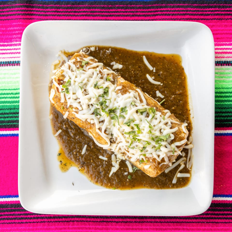 A golden-brown enchilada covered in shredded white cheese and cilantro, swimming in green salsa.