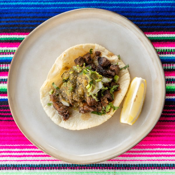 A single carne asada taco with green salsa, onions, and cilantro, served with a lemon wedge.
