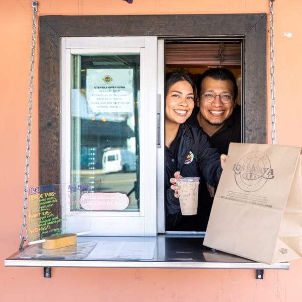 Two smiling restaurant employees hand out a drink and a branded paper bag from a takeout window.
