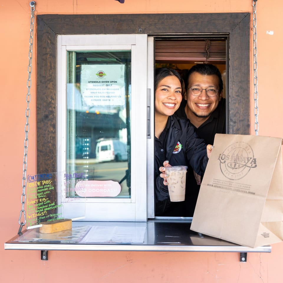 Two smiling restaurant employees hand out a drink and a branded paper bag from a takeout window.