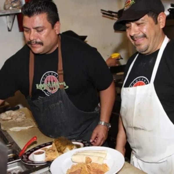 Two male chefs in aprons prepare food in a kitchen, with plates of tamales and chips in the foreground.