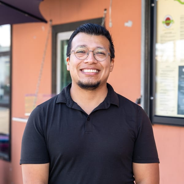 Smiling man in glasses and a black polo shirt standing outside a restaurant.