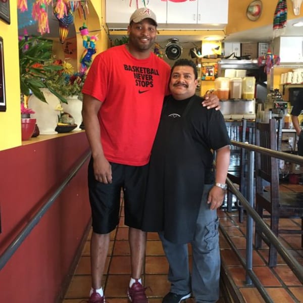 A tall man in a red shirt and a shorter man in a black apron pose together in a colorful restaurant.