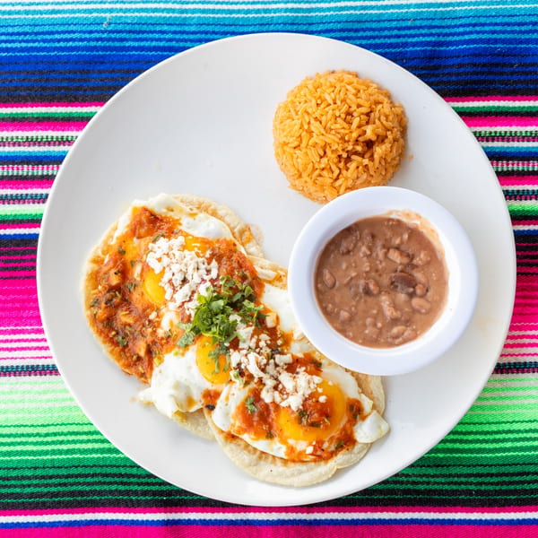Huevos rancheros with rice and beans on a white plate over a colorful striped tablecloth.
