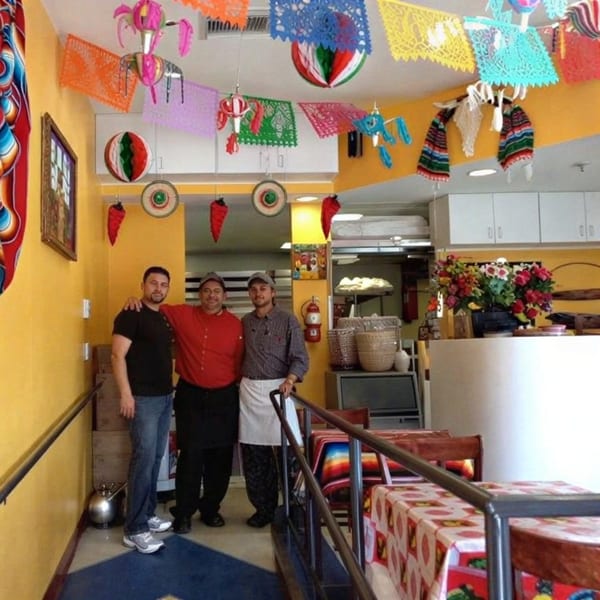 Three male restaurant staff members stand together in a brightly decorated Mexican restaurant.