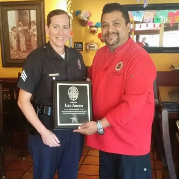 A police officer and a chef in a red uniform smiling and holding a plaque together.